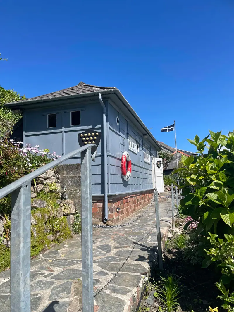 The blue-painted cabin with a red-and-white lifebuoy on its wall, beside a flagstone garden path under a clear blue sky