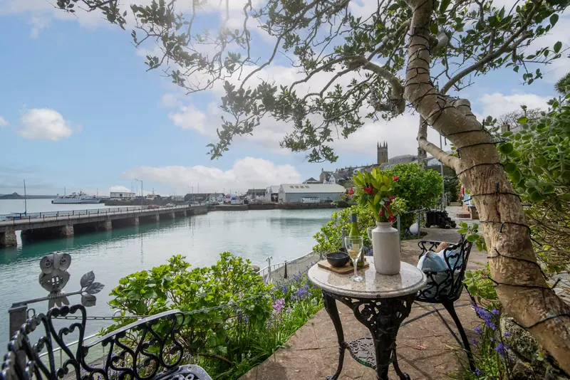 A garden table set with glasses and flowers beneath a tree, overlooking Penzance Harbour
