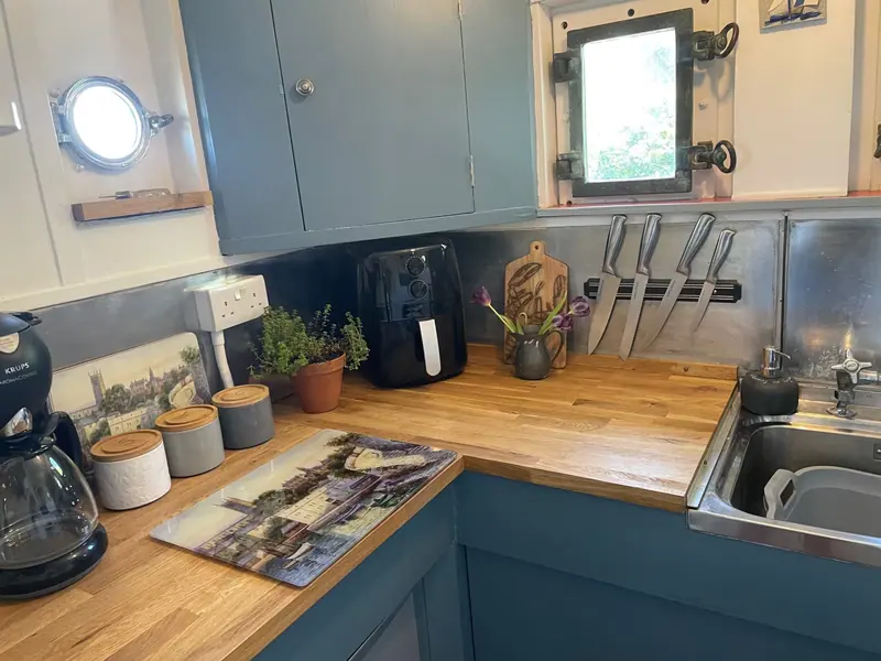 The kitchen, with blue-grey cupboards, wooden worktops, a small porthole, a coffee machine and a steel sink