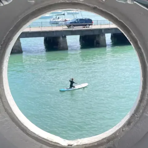 A paddleboarder on the green water of the harbour, seen through one of the cabin’s round portholes