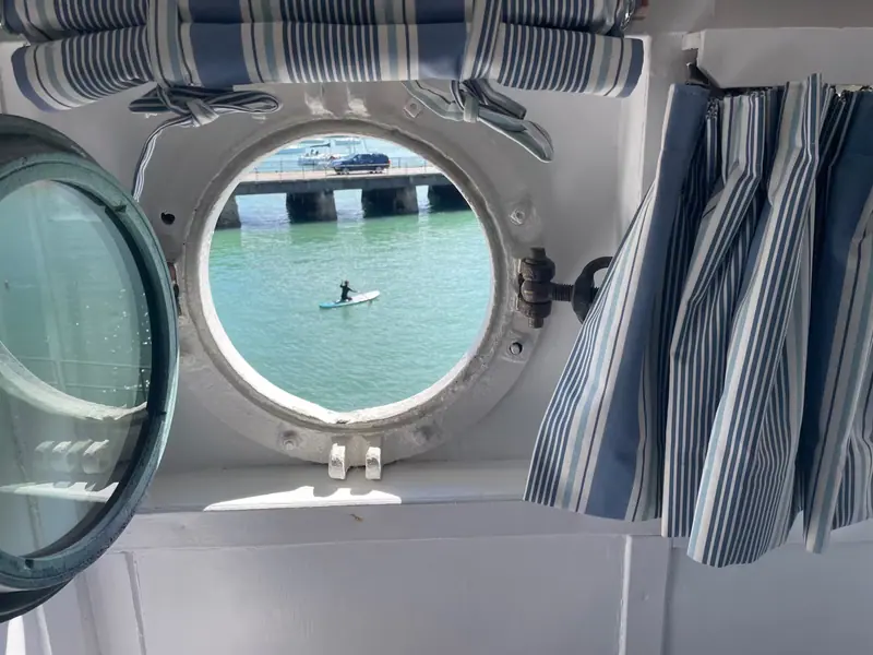 A paddleboarder on the harbour, seen through a round porthole framed by blue-and-white striped curtains